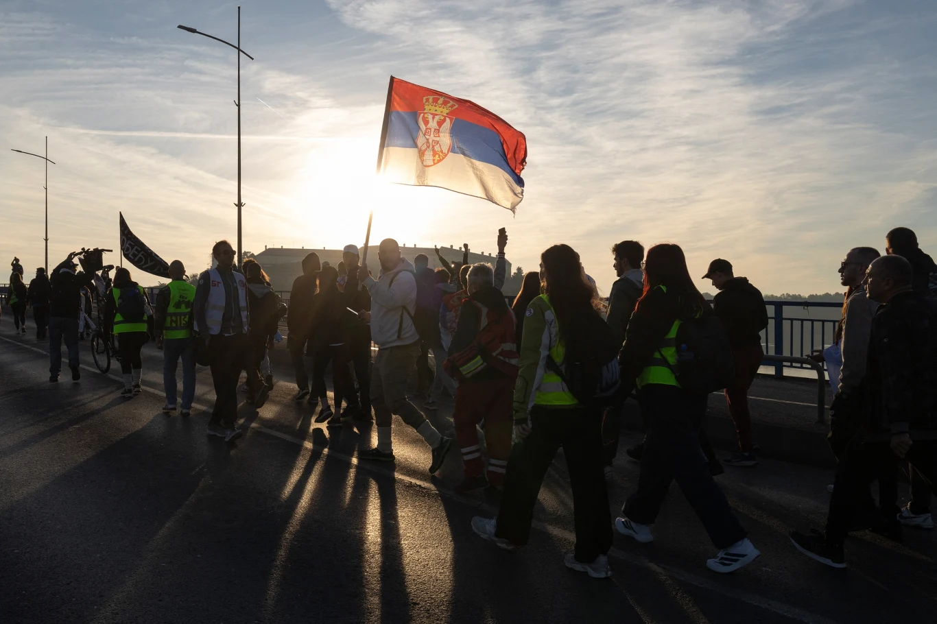 Protesty studentów w Nowym Sadzie, Serbia Grupa ludzi maszerujących ulicą z dużą serbską flagą niesioną przez jednego z uczestników, silnie podświetleni przez zachodzące słońce na tle miasta.