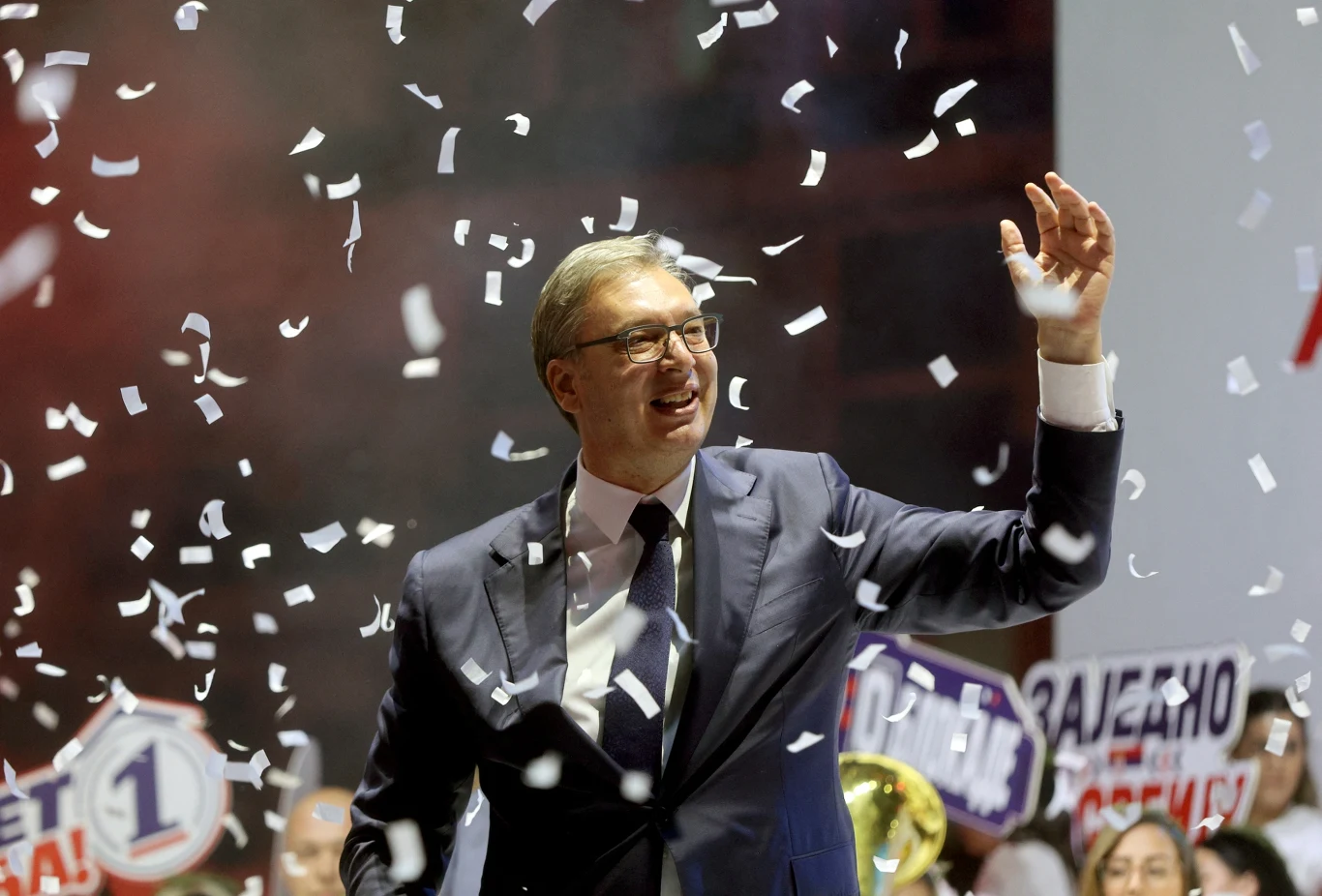 Aleksandar Vučić podczas kampanii wyborczej Mężczyzna w garniturze uśmiecha się i macha ręką podczas ulicznej uroczystości, otoczony opadającym konfetti i tłumem osób trzymających transparenty wyborcze.