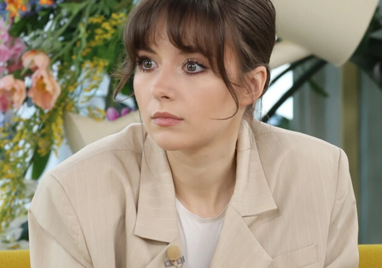 Kasia Sienkiewicz Kasia Sienkiewicz w beżowej marynarce siedzi na żółtej kanapie w otoczeniu kwiatów i roślin w studiu telewizyjnym.