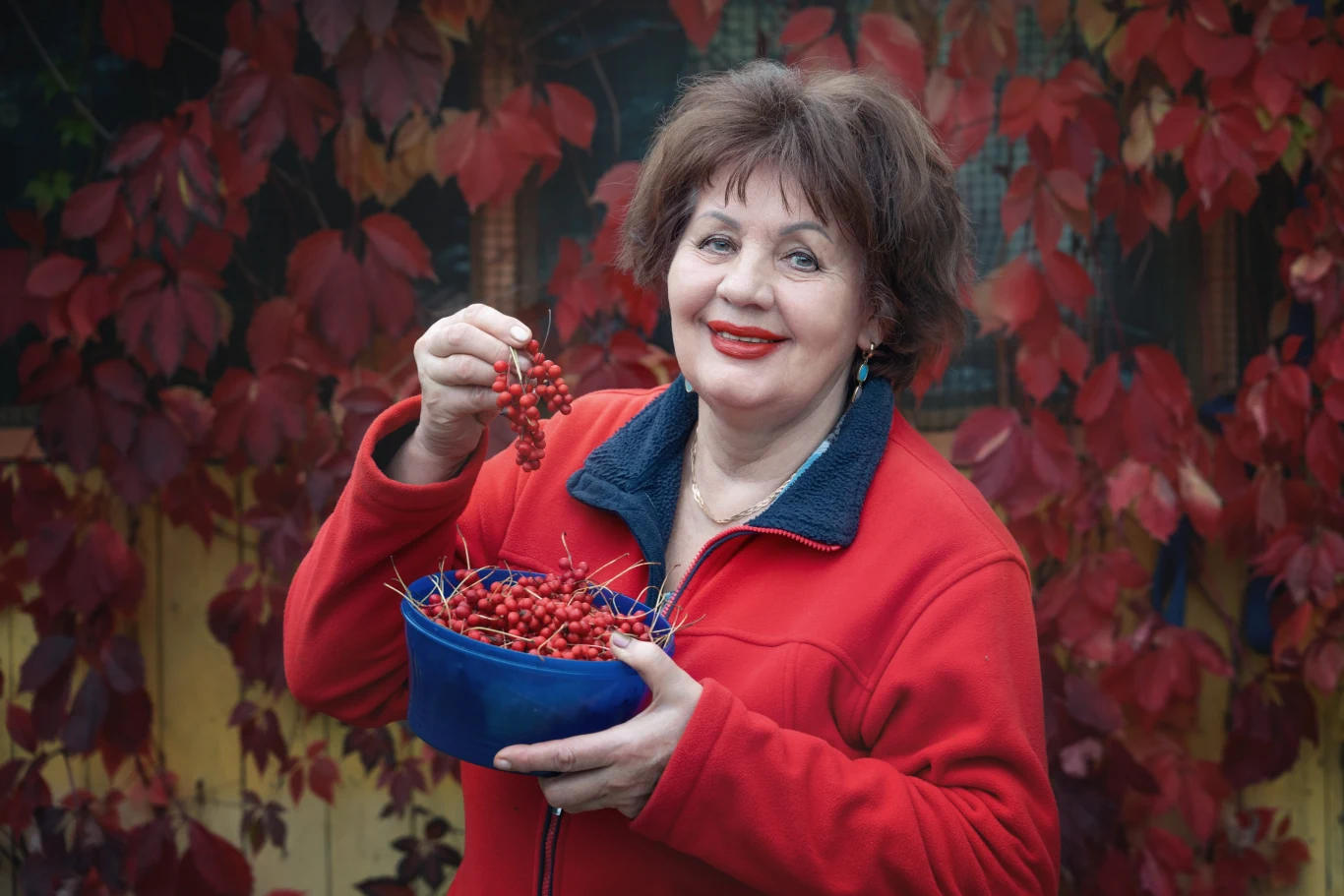 Imię Kalina zyskuje na popularności. Kojarzy się z jesienią. Kobieta w czerwonej kurtce trzymająca miseczkę oraz podnosząca do góry kiść czerwonych owoców, na tle czerwonych liści.