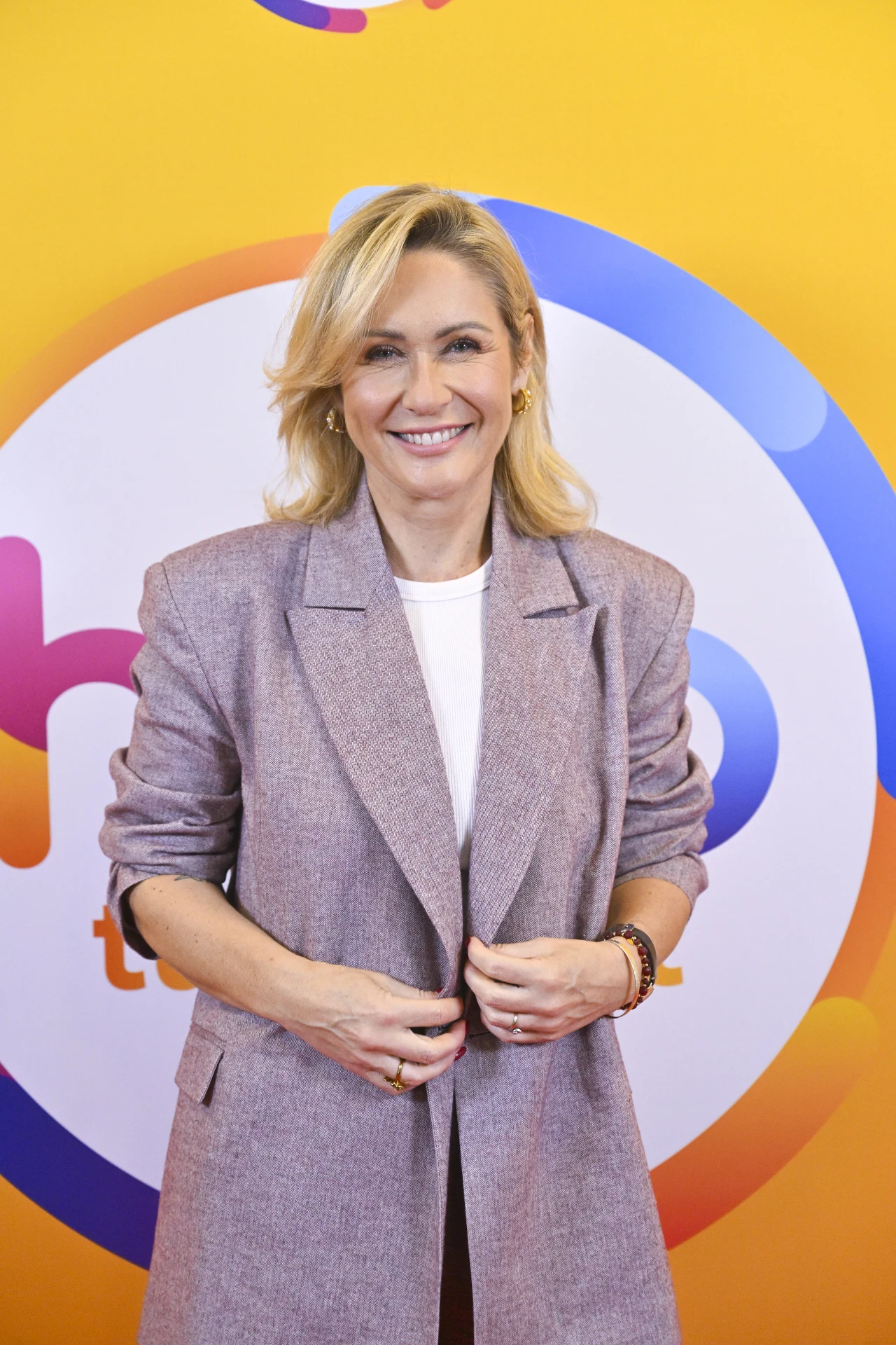 Uśmiechnięta kobieta o blond włosach w jasnej marynarce, stojąca na tle kolorowej grafiki z okrągłym logo.
