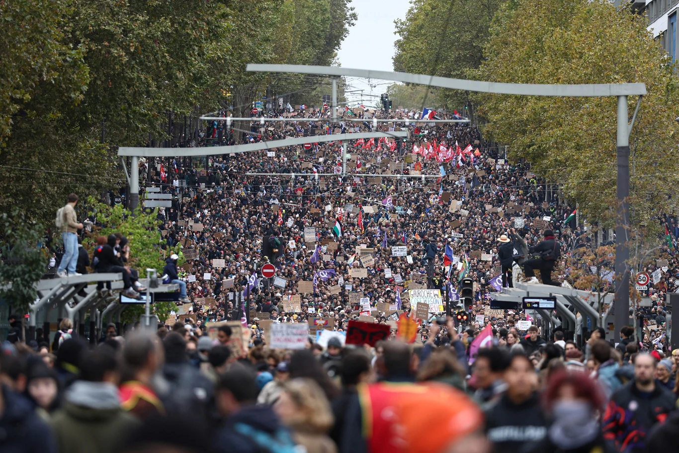 Wielka demonstracja w Paryżu we Francji Tłum ludzi maszerujących główną ulicą między drzewami, wiele osób trzyma transparenty i flagi, atmosfera wyraźnie manifestacyjna.