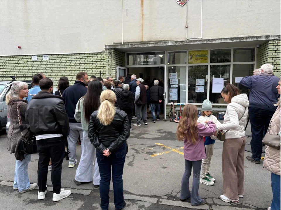 Kolejka ludzi ustawiająca się przed wejściem do budynku, w którym na drzwiach widoczne są informacje oraz plakaty; część osób to rodziny z dziećmi, większość patrzy w stronę drzwi.