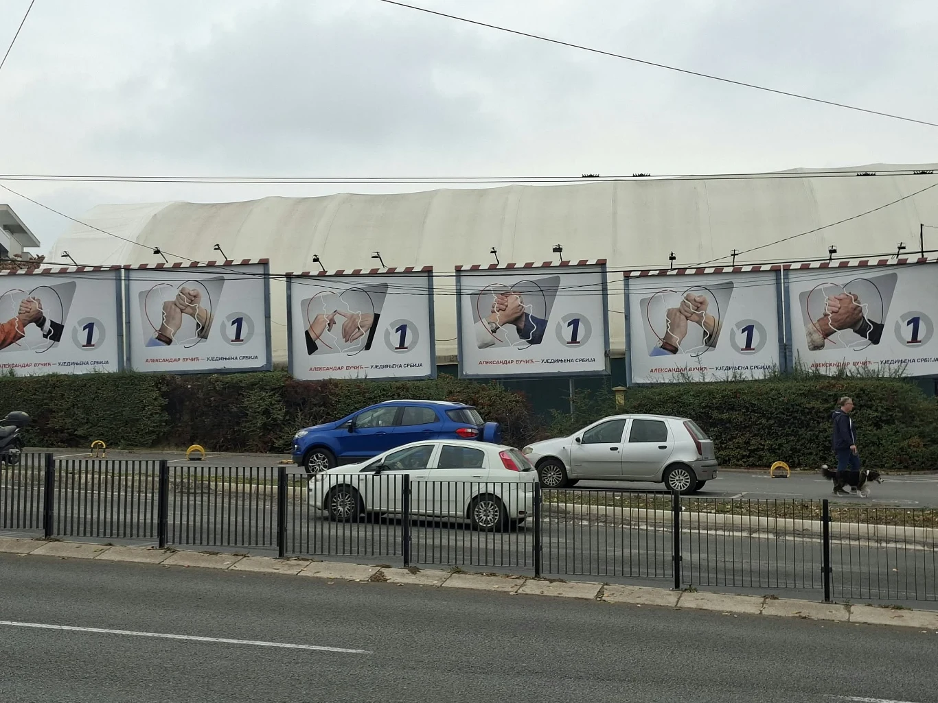Rząd identycznych billboardów z wizerunkiem starszego mężczyzny w okularach i białym stroju, który przykłada rękę do ucha, stojący za rzędem samochodów zaparkowanych przy ulicy.