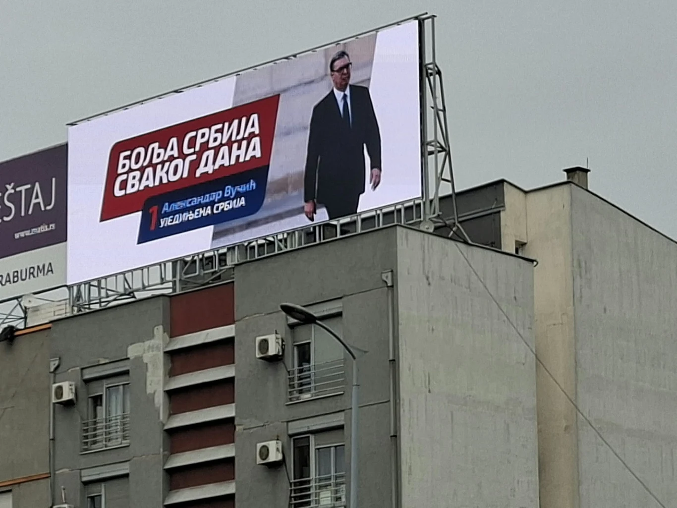 Na dachach, na słupach, na billboardach. Aleksandara Vučicia pełno w Belgradzie Duży billboard z wizerunkiem mężczyzny w garniturze oraz tekstem w języku serbskim umieszczony na dachu wielopiętrowego budynku mieszkalnego.