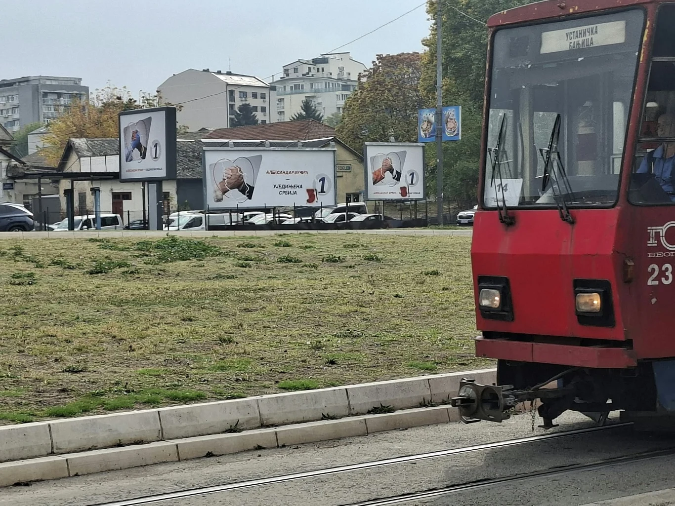 W stolicy Serbii pełno wyborczych billboardów - zwłaszcza tych wywieszonych przez SNS Czerwony tramwaj jadący wzdłuż torów na tle trzech dużych billboardów reklamowych oraz kilkupiętrowych budynków mieszkalnych w oddali.