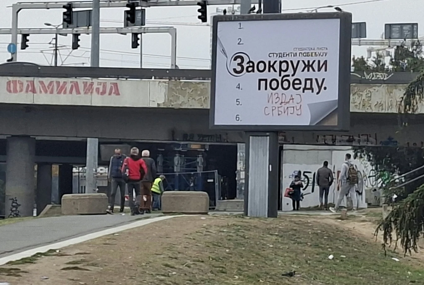 Zdewastowany billboard Listy Studenckiej Grupa osób idących chodnikiem pod wiaduktem, obok dużego billboardu z napisem w cyrylicy, na którym widnieje lista oraz hasło wyborcze, w tle widoczne elementy miejskiej infrastruktury i graffiti.