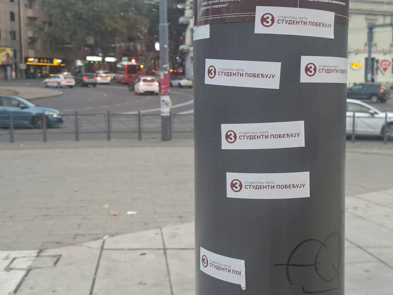 Wlepki promujące Listę Studencką - Studenci Wygrywają Kilka naklejek z napisem w cyrylicy i liczbą 3 przyklejonych do szarego słupa na tle miejskiej ulicy z samochodami i budynkami.
