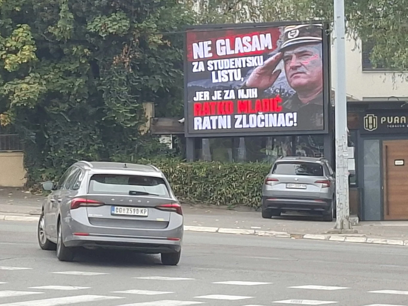 Wyborcza grafika dotycząca Ratko Mladicia, uderzająca w Listę Studencką Billboard z wizerunkiem mężczyzny w mundurze wojskowym oraz dużym, kontrastowym napisem w języku serbskim i dwoma zaparkowanymi samochodami na ulicy.