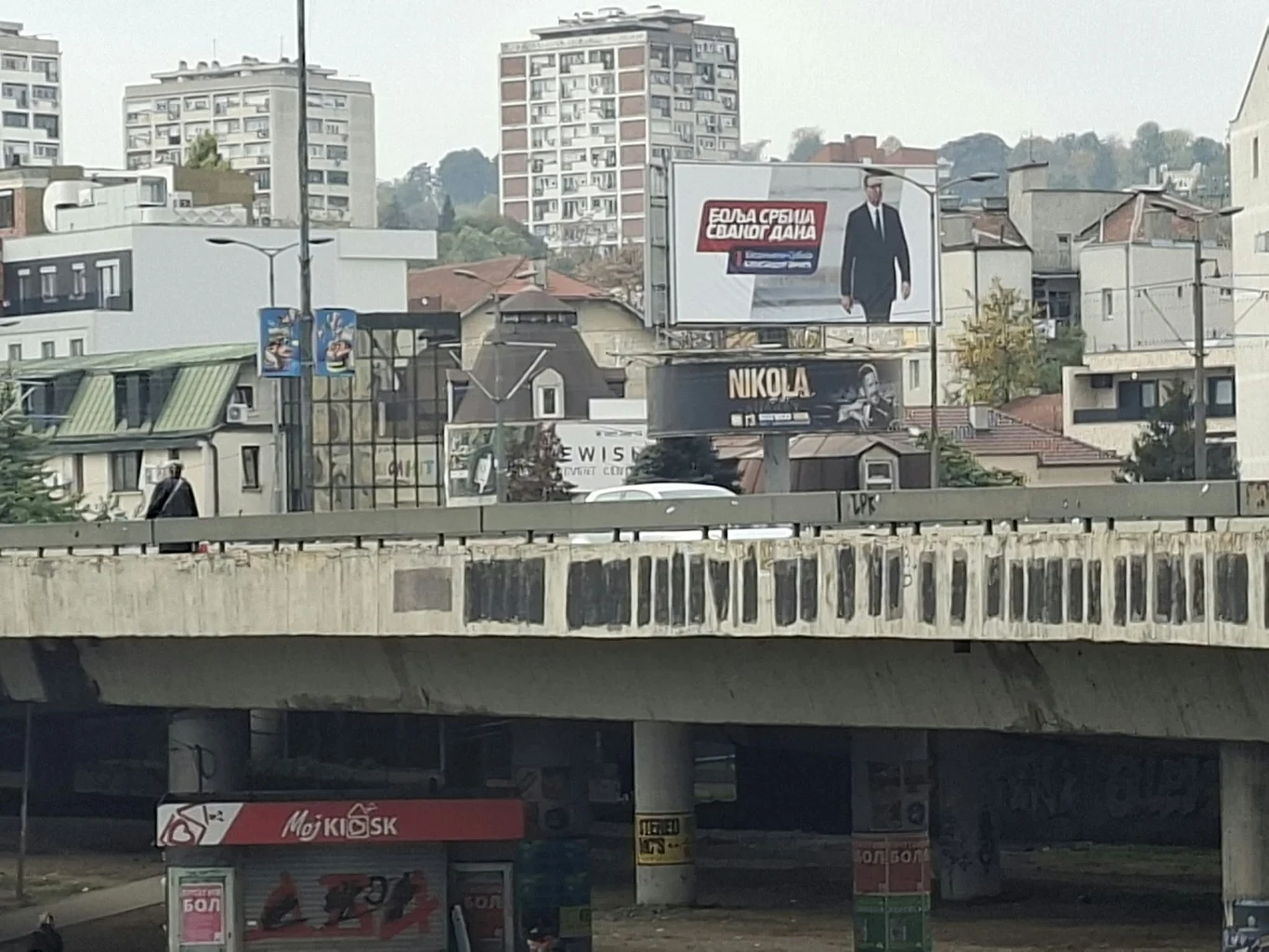 Jeden z billboardów Vučicia przy ważnym węźle drogowym w Belgradzie Duży billboard z sylwetką mężczyzny w garniturze dominuje nad miejskim krajobrazem, w tle widoczne są bloki mieszkalne i budynki mieszkalne, a na pierwszym planie znajduje się kiosk oraz częściowo zniszczony most.