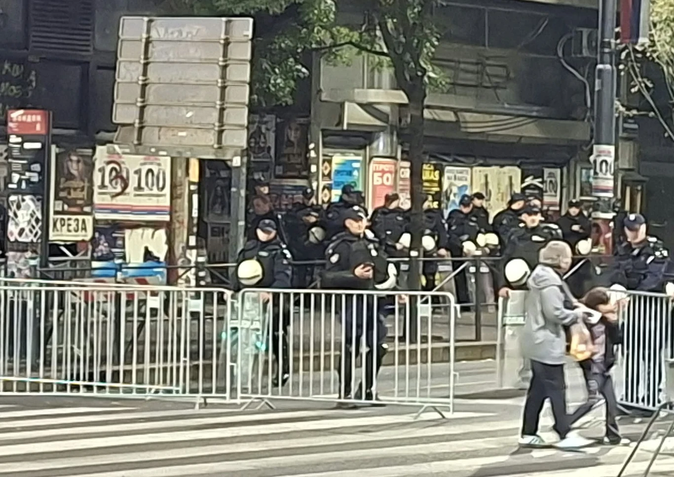 Policja zamknęła dla ruchu niektóre ulice w centrum Belgradu Grupa policjantów wyposażonych w kaski i mundury ochronne stojących za metalowymi barierkami ustawionymi przy ulicy, z przodu dwie osoby przechodzą przez przejście dla pieszych.