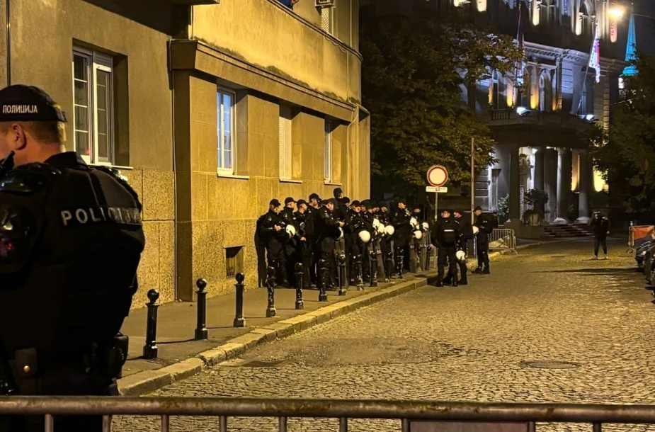 Grupa policjantów w uniformach i kaskach ochronnych ustawionych w szeregu wzdłuż budynku przy oświetlonej ulicy nocą.