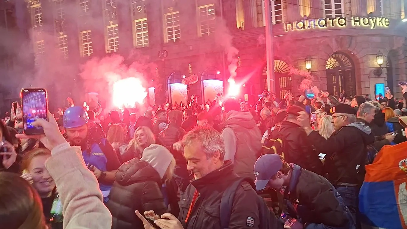 Feta na ulicach Belgradu Tłum ludzi zgromadzonych na ulicy przed podświetlonym budynkiem, kilku uczestników trzyma race emitujące czerwone światło i dym, część osób rejestruje wydarzenie telefonami.