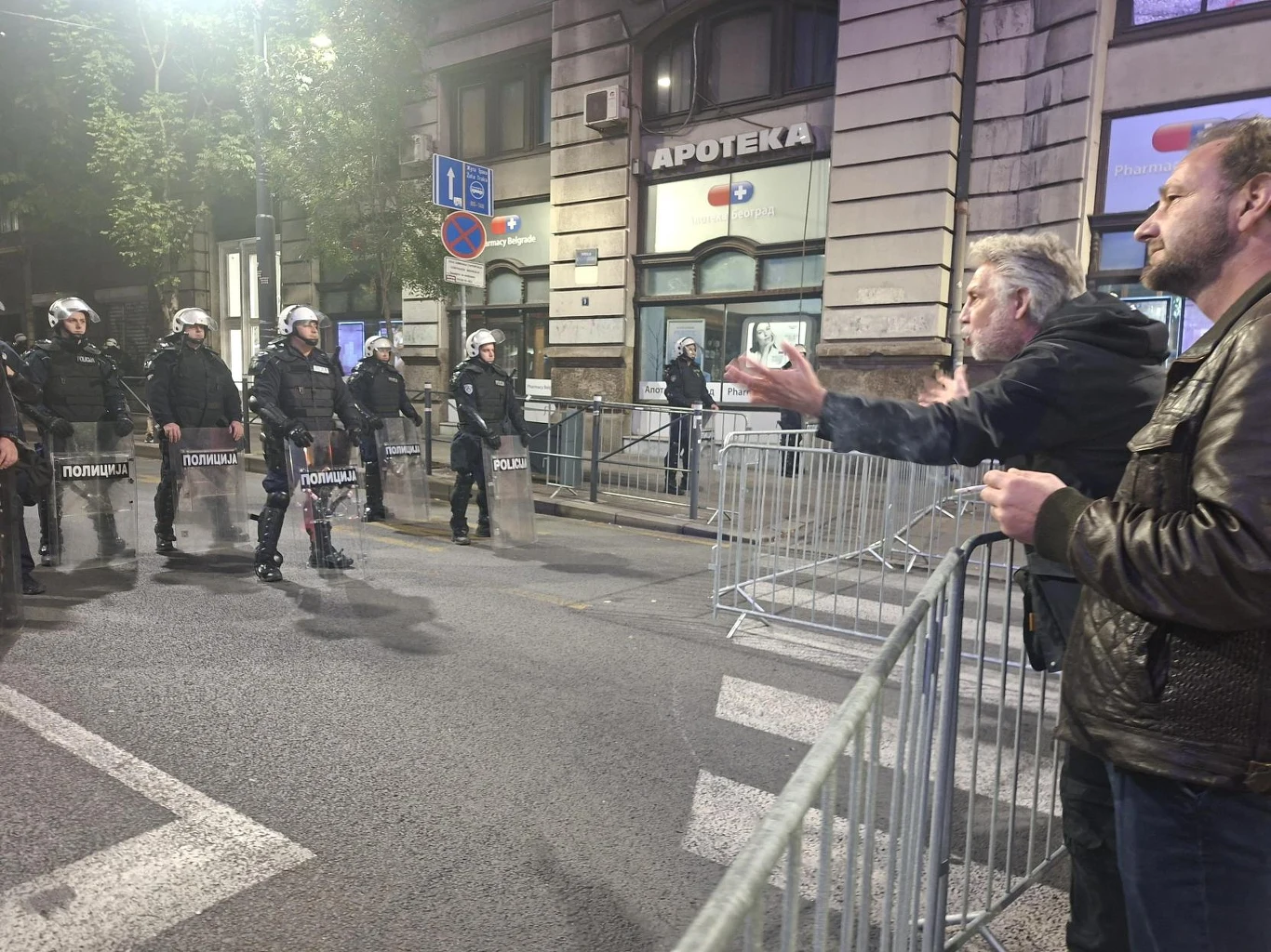 Policja na ulicach Belgradu Grupa policjantów w pełnym umundurowaniu i z tarczami zabezpiecza ulicę, naprzeciwko stoją dwaj mężczyźni gestykulujący za metalowymi barierkami, tło stanowi fasada miejskiego budynku z widocznym szyldem apteki.