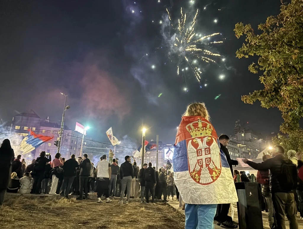 Fajerwerki na Placu Slavija Tłum ludzi zgromadzony w centrum miasta, jedna osoba owinięta flagą Serbii, kolorowe fajerwerki na nocnym niebie, wokół powiewają serbskie flagi.