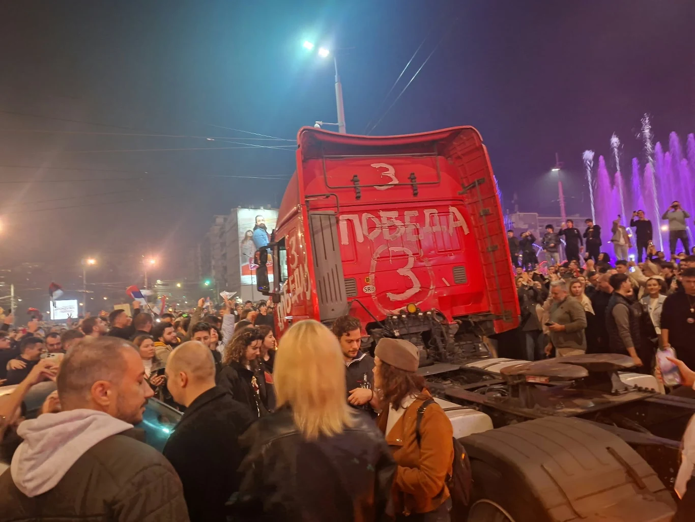 Na tirze znalazły się hasła wsparcia dla listy numer 3, czyli Listy Studentów Tłum ludzi zgromadzony wokół czerwonej ciężarówki z napisem w cyrylicy, która stoi na ulicy w pobliżu podświetlonych na fioletowo fontann.