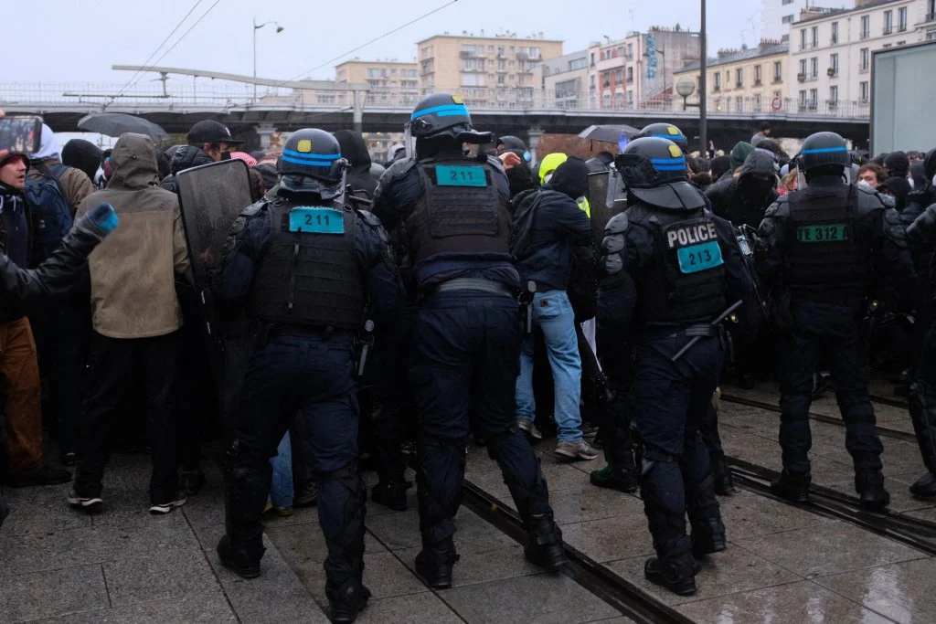 Ustawieni w szeregu funkcjonariusze policji w pełnym wyposażeniu ochronnym próbują powstrzymać tłum ludzi na ulicy w miejskiej scenerii.