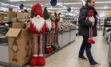 Mężczyzna w grafitowej parce niesie pod pachą czerwonego skrzata na długich pasiastych nogach obok wyspy z kartonem i rzędem skrzatów, misiem i reniferem w dyskoncie 