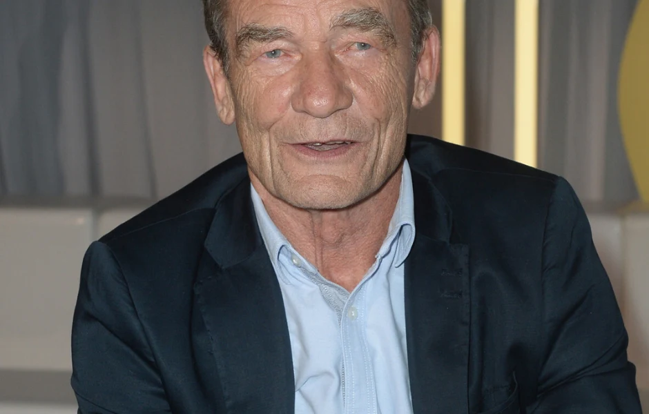 Krzysztof Kiersznowski Mężczyzna w średnim wieku ubrany w granatową marynarkę i jasnoniebieską koszulę, siedzący we wnętrzu o neutralnym tle.