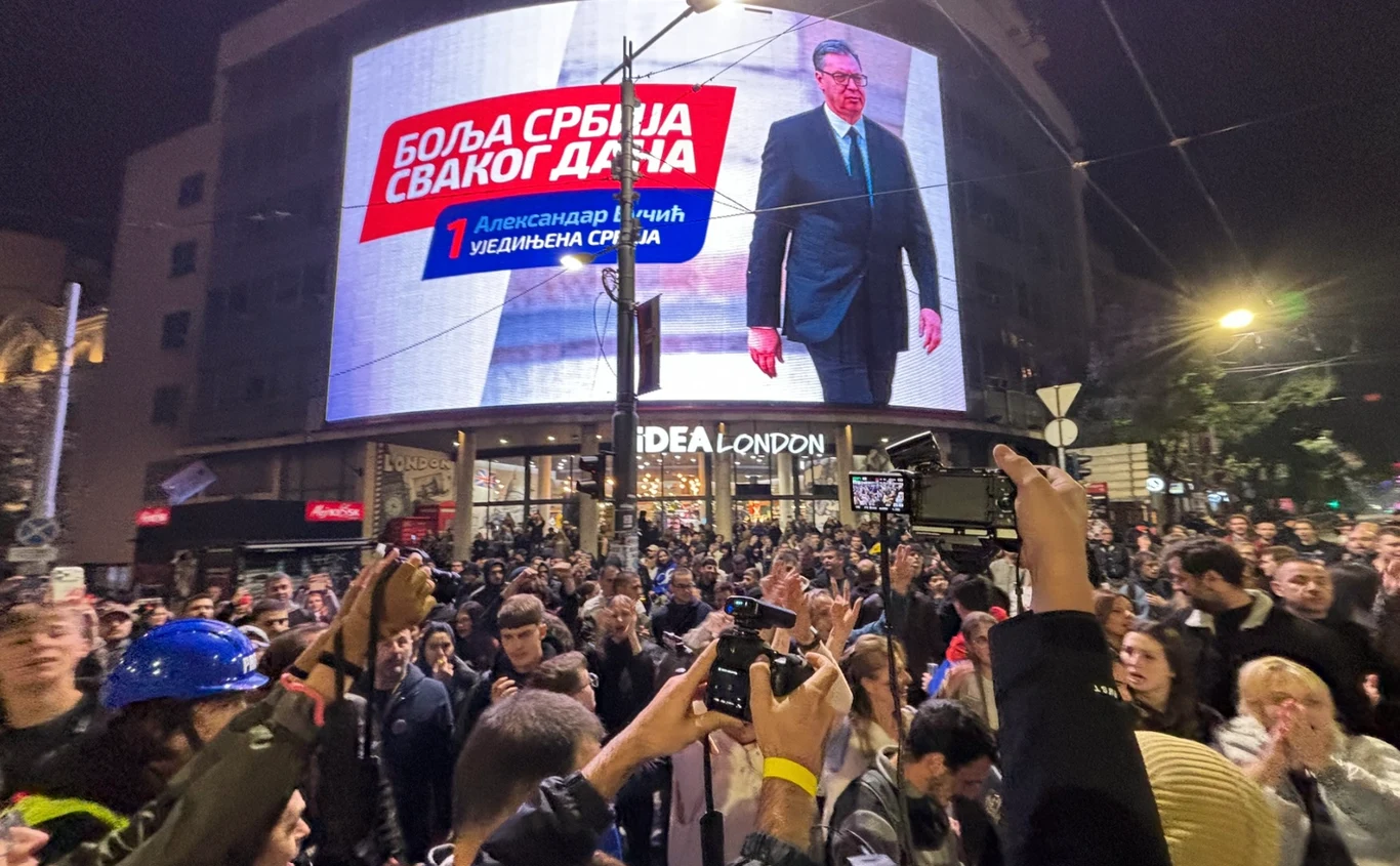 Zwolennicy Listy Studenckiej przed materiałem wyborczym Vučicia tuż po ogłoszeniu wyników wyborów Duży tłum ludzi zgromadzony pod wielkoformatowym ekranem prezentującym sylwetkę mężczyzny w garniturze, nad którym widnieje napis w języku serbskim, większość osób trzyma telefony w dłoniach i rejestruje wydarzenie.