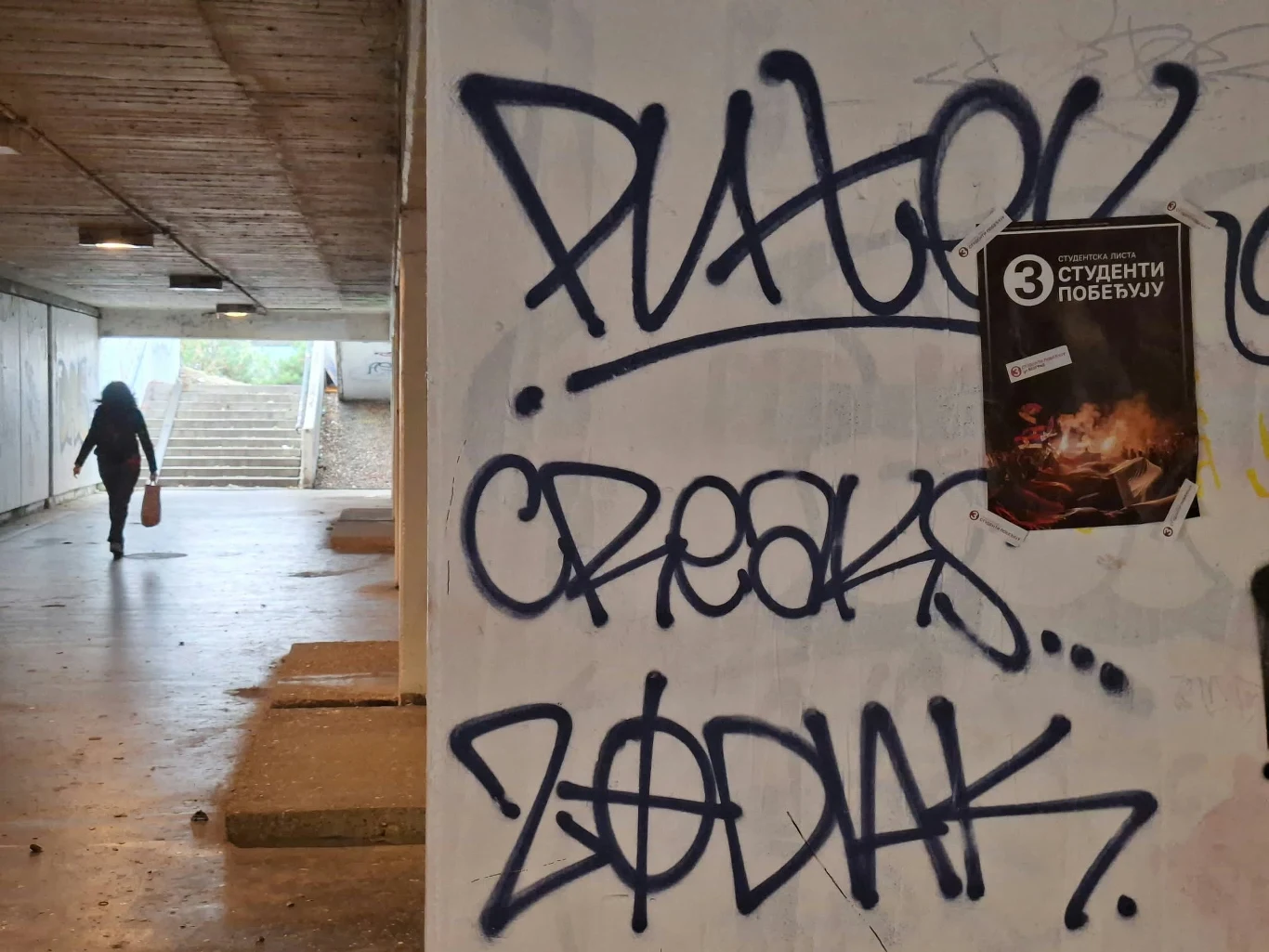 Duże napisy graffiti wykonane niebieskim sprayem na białej ścianie tunelu, w tle osoba z torbą idąca w stronę schodów prowadzących na zewnątrz.