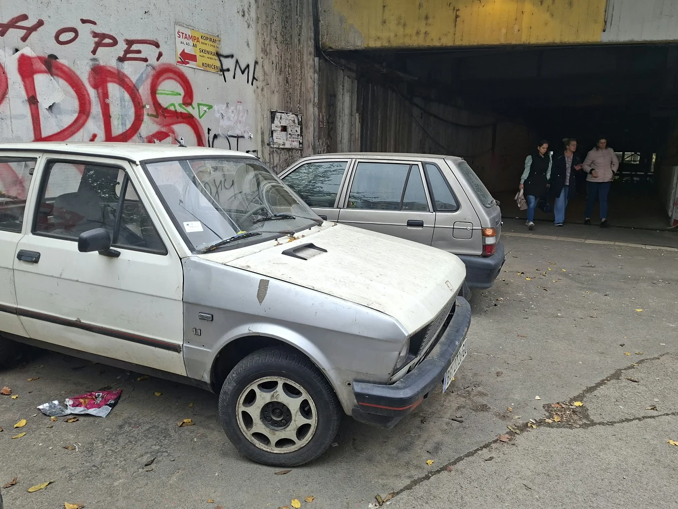 Dwa stare, zaniedbane samochody osobowe zaparkowane przed wjazdem do ciemnego tunelu, obok ściany pokrytej kolorowymi graffiti, w tle kilku przechodniów wchodzących do tunelu.