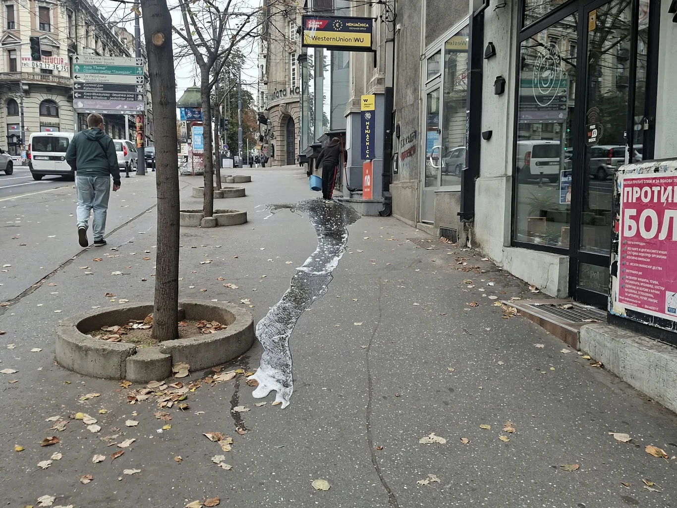 Chodnik miejski z widocznym jasnym śladem po rozlanej cieczy biegnącym wzdłuż krawędzi, po lewej stronie rządz drzewa w betonowych donicach, po prawej fasada budynku z witryną i reklamą, w tle kilka osób idących ulicą.