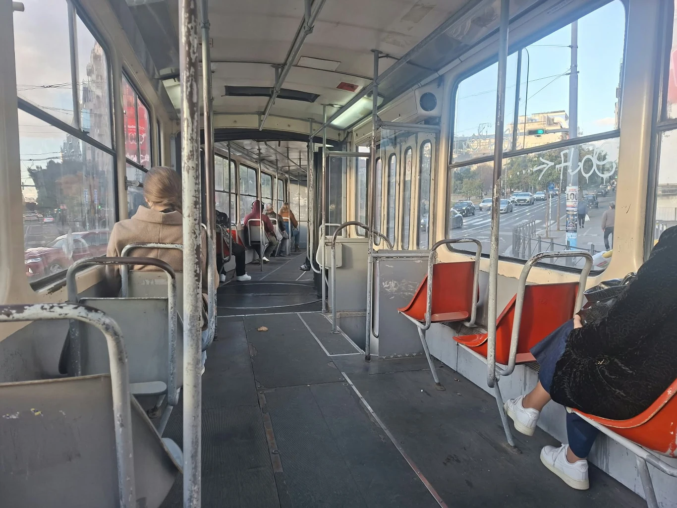 Wnętrze starego tramwaju z czerwonymi siedzeniami, kilku pasażerów siedzi w przestronnej części pojazdu, przez okna widoczne miejskie zabudowania.
