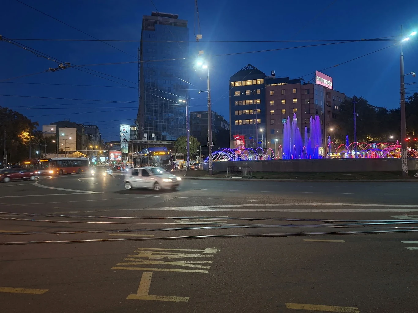 Oświetlona fontanna z kolorowymi światłami w centrum ruchliwego skrzyżowania, otoczona wieżowcami i przejeżdżającymi samochodami po zmroku.