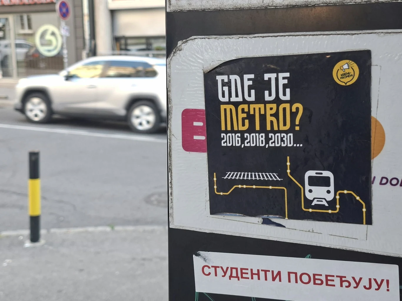 Plakat z napisem 'GDE JE METRO? 2016, 2018, 2030...' oraz grafiką symbolizującą linię metra i ikonę pociągu, umieszczony na słupie przy ruchliwej ulicy, obok naklejki z napisem 'СТУДЕНТИ ПОБЕЂУЈУ!'