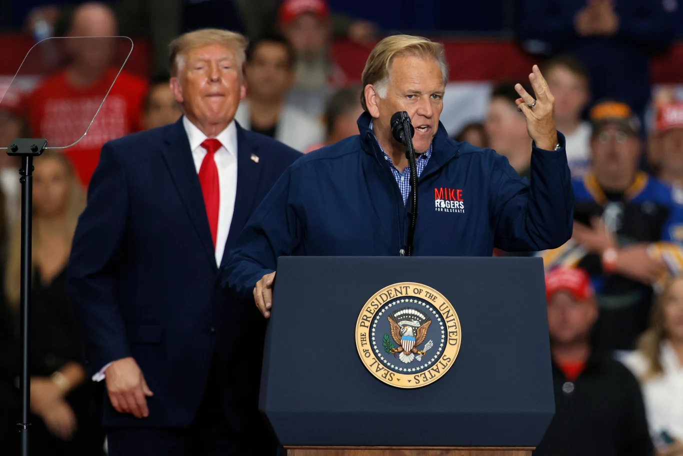 Mike Rogers dostał wsparcie Donalda Trumpa na wiecu w Michigan Mężczyzna przemawia za mównicą oznaczoną pieczęcią prezydenta Stanów Zjednoczonych, za nim stoi inny mężczyzna w garniturze, a w tle widoczny jest tłum zgromadzonych osób.