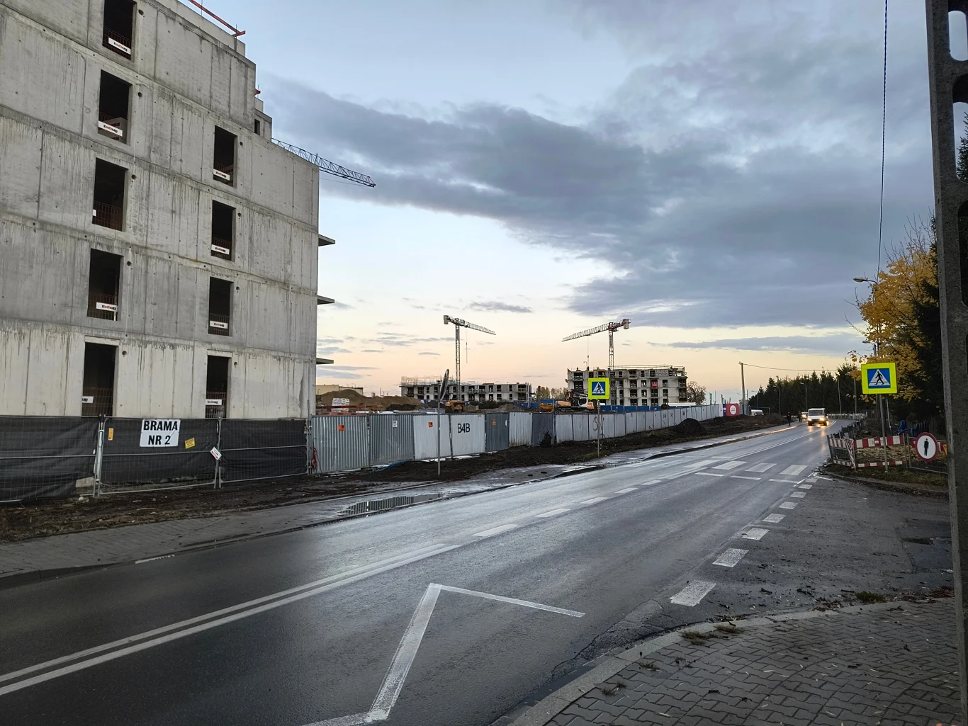 Piękne osiedle bez drzew Nowy budynek mieszkalny w stanie surowym po lewej stronie ulicy, ogrodzenie budowy oraz żurawie wieżowe unoszące się nad kolejnymi etapami inwestycji po drugiej stronie drogi, asfaltowa jezdnia z oznakowaniem poziomym i znakiem przejścia dla pieszych.