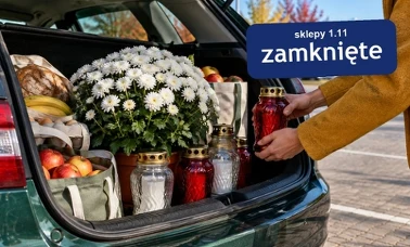 Otwarty bagażnik auta na parkingu z doniczką białych chryzantem, szklanymi zniczami i torbami z zakupami, do którego dłonie wkładają czerwony znicz 
