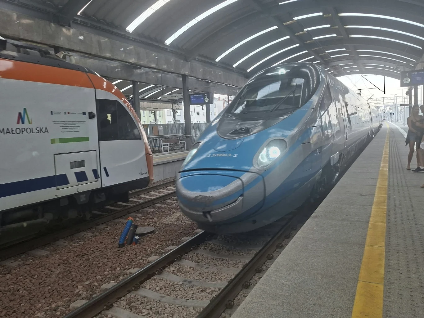 Dwa nowoczesne pociągi zatrzymane przy peronie na zadaszonej stacji kolejowej, jeden w biało-niebieskim malowaniu, drugi w barwach pomarańczowo-białych z widocznym napisem Małopolska.