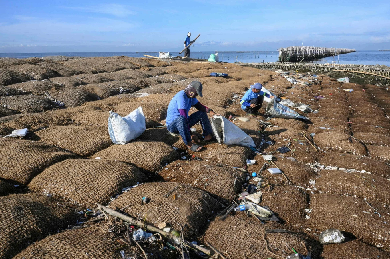 Kilka osób w odzieży ochronnej zbierających śmieci z dużych worków ułożonych na rozległej plaży przy brzegu morza, wokół rozrzucone odpady i plastikowe butelki.