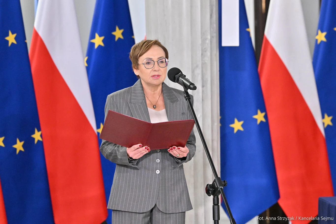 Sędzia Anna Korwin-Piotrowska w garniturze przy mikrofonie, na tle flag Polski i Unii Europejskiej.