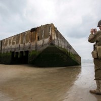 Jeden z pasjonatów historii II wojny światowej, ubrany w replikę mundur wojskowego z czasów wojny, fotografujący wrak pontonu na plaży w Arromanches